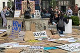 A photograph of the empty pedestal where the statue of Edward Colston once stood, taken on 7 June 2020. The pedestal has graffiti art sprayed on it, and surrounding it are Black Lives Matter and antiracist protest signs. In the background, a small crowd of people are gathered, with a couple of people taking a picture of a Black man beside the empty pedestal with a raised fist and holding a sign in the other hand.