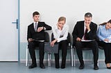 A group of 4 professionally dressed people looking bored sitting outside an office door while waiting to interview.