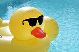 Large, yellow duck-shaped inflatable with red beak and black sunglasses sitting in a swimming pool.