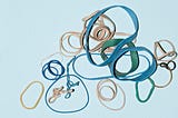 Lots of different circular rubber bands on a bright background thrown on top of each other. Some are twisted, others are nearly round.