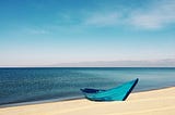 A turquoise row boat washed ashore on a deserted sand beach with a calm turquoise ocean and sky in the background