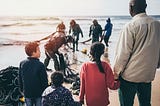 The front part of the image shows a group of people look afar, comprised of an adult and three children. They are holding hands, which suggests that they are a family. At the back, other individuals are seeing picking things from a fishnet at the seashore.