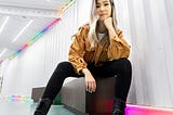 A young woman with long, silver hair sits on a bench looking down at the camera. She’s wearing black jeans and a camel colored jacket with black boots. Strips of neon lights border the room behind her.