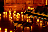Grand piano and cushioned bench surrounded by glowing candles in a warmly lit, wood-panelled concert hall.