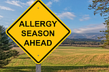 Yellow sign in front of field reading “Allergy Seasons Ahead”