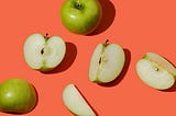 A photograph of several green apples, some cut apart with the cut side facing up, scattered across a bright, almost neon red-pink background.
