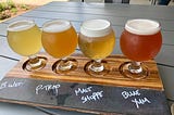 A flight of four beers on a wooden tray on an outdoor table, with labels containing the name of each beer.