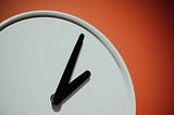 Photo of blank white clock face with black minute and hour hands pointing to roughly 2 p.m. against an orange-red background.