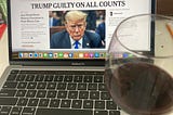 A glass of red wine sits on the edge of a laptop with The New York Times open to a story “TRUMP GUILTY ON ALL COUNTS.” Books, a pencil are also seen on the white desk. (Photo by author.)