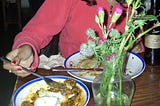 Photo showing a dinner table with two plates of half eaten food. A vase of flowers are on the table and a person opposite in a pink hoodie is reaching for a plate with their fork. The image is cropped so their face is not visible.