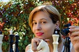 An attractive young woman playing chess in the open air. She is holding a black knight.