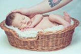 A human baby sleeping in a straw basket, with a hand of an adult caressing their head.