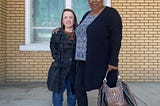 A redhead dwarf woman stands next to an average height African-American woman in front of a yellow brick church