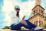 A photo of a woman listening to a podcast on her phone with the Eiffel tower (Paris) in the background.