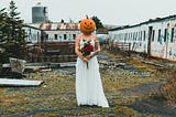 a woman with a pumpkin head wearing a white off-shoulder dress and holding a red and yellow flower bouquest is standing in a discarded place with rows of white boarded up house behind her