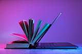 Livro aberto encostado em um laptop semiaberto, iluminados por luzes coloridas em tons de rosa e azul, sobre uma superfície plana.