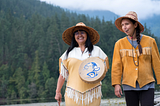 Two Indigenous Canadian women wearing traditional clothing walk by a lake. One woman is holding a hand drum. They are both happy and smiling.