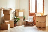 Cardboard moving boxes stacked in a sunlit room with plants and a wooden door.