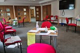 A room with red chairs around the outside of it, some of which have pieces of paper on them. There are three tables that have papers, cleaning spray and bottles of hand sanitizer on them.