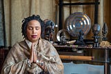 photos Aishah, brown skinned Black woman, wrapped in a golden shawl, with palms together, and eyes closed. She is seated in front of an altar with statues of West African Deities, Buddha, Quan Yin, and a gong.