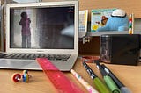 A laptop on a desk with a photo of a child in a storm, yelling on the desktop. There are pens and pencils on the desk, a stim toy and a ruler. In the back is a French book about winter and a stuffed Among Us video game character.