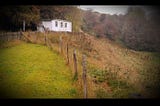 In the top left corner of the picture there is a white cottage on a hill with green and brown fields all around. In the top right corner there are some trees in the brown colours of autumn. Bottom centre of the picture some wooden polls and wire fence