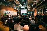 Chuck speaking with a microphone, presenting Design Sprints on a projector to a live audience at WeWork Spinningfields, Manchester. Room is softly lit, and has an industrial style.