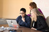 three employees collaborate around a laptop computer sitting on a table