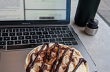 Lauren’s computer, the screen blurred, with a lovely looking mocha coffee in front of it, complete with marshmallows and chocolate drizzle. To the back right of the photo sits a green water bottle.