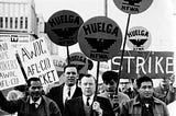 Black and white photo of Larry Itliong, Cesar Chavez and others holding signs that say, “Huelga NFWA,” “strike,” and “AWOC AFL-CIO Picket” during the Delano Grape Strike of 1965