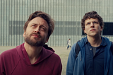 Jesse Eisenberg and Kieran Culkin look upward with serious expressions in front of the POLIN Museum’s glass-panel façade in A Real Pain (2024).