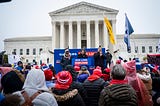 A crowd gathers in front of the Supreme Court for a rally. Signs and banners say, “gun laws save lives”