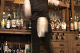 A bartender standing on a back bar about to pick a bottle of spirits
