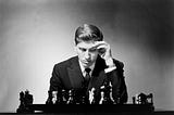 In the center of the composition is a focused young man, Bobby Fischer. His head is bent as he gazes at the chessboard. His left hand touches his forehead, emphasizing the chess player’s concentration. The photo is black and white.