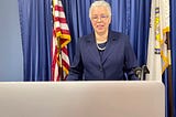 Cook County Board President Toni Preckwinkle stands behind a podium delivering a virtual keynote address to the League of Women Voters of Cook County. She is wearing her glasses, a pearl necklace, and navy blue shirt and blazer. On the sides are the American Flag and the Cook County flag. A laptop can also be seen.