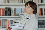 Woman wearing white with short dark hair holds a pile of books that reach her chin. She stands in front of white bookshelves lined with books and she is looking at the camera.