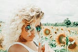 A woman with curly blonde hair poses in a sunflower field, wearing sunglasses and a playful expression.