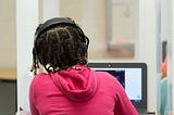 A girl in a pick sweatshirt wears headphones and types on her laptop while seated at a desk behind a protective screen