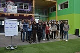 A group of people stand inside a building with green walls and turf flooring; a large white paper easel stands on the left side of the image with purple handwritten words on it saying “MIT Media Lab x Smart Living Lab.”