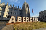 A large sign saying “Aberdeen” is written in large letters on a patch of grass. Behind it is a large building with Gothic architecture. The sky is blue.