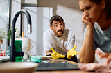 A frustrated man wearing yellow rubber gloves gestures emphatically while standing at a kitchen sink, with green dish soap and cleaning supplies nearby. In the foreground, a woman sits with her head resting on her hand, looking upset and disengaged, highlighting tension in the scene