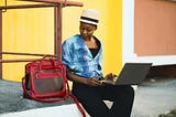 Woman sitting in front of a yellow wall beside a red bag while using a laptop