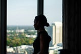 A woman is standing by a window in what appears to be an office building. She is looking out thoughtfully.