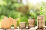 Wooden carved house sitting to the left of three progressively taller stacks of money coins with green leaves sprouting