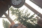 Sarah looks up at a vintage clock outside under a high verandah. Fir trees are in the background. Original photo by Sarah Smith © 23 Oct 2016