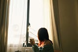 A woman sits beside a sunlit window holding a warm drink as soft morning light fills the room, conveying peace, silence, and contemplative presence.