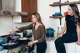 two female friends in a kitchen. One is standing at the stove, cooking. the other is sitting on the counter behind her, looking on.