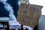 A protest sign, written on cardboard, reading “Stop asking me to forgive fascism.”