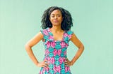 A Black woman with dreads wearing a teal-and-pink floral dress. She has a confident posture — hands on her hips, chest out. She has a smirk on her face. The picture was taken in a light-blue background.