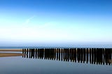 Landscape of Burgh-Haamstede, in the Netherlands, depicting a sandy beach with sea in the distance and shallow water in the foreground. There is a line of posts — wave breakers — driven into the sand.